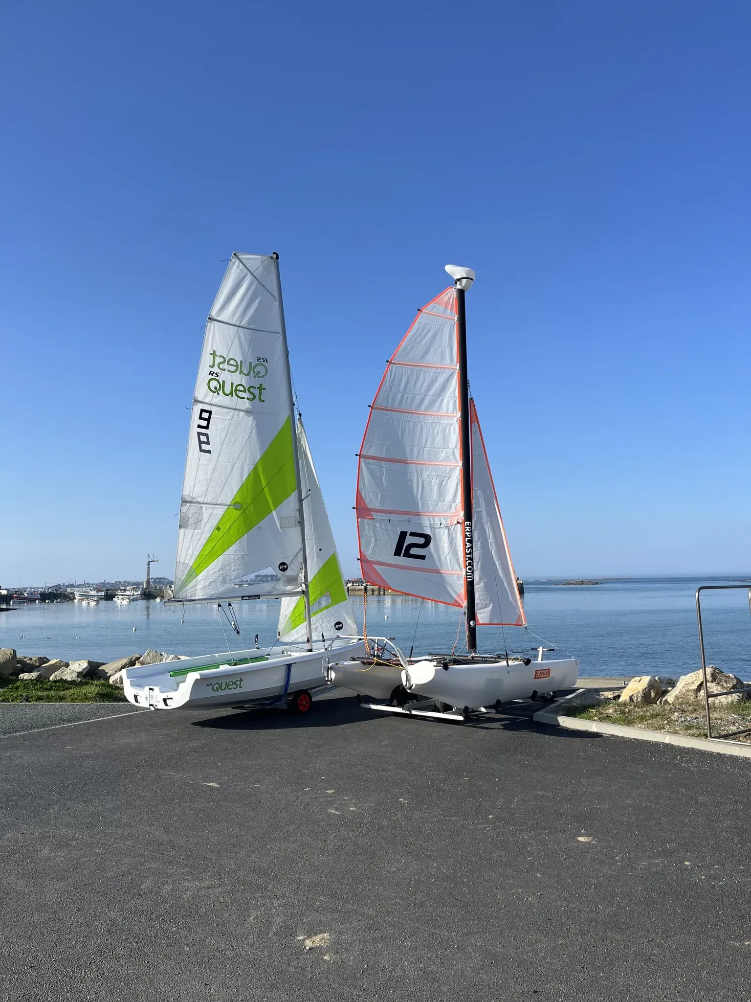 Un dériveur RS Quest et un catamaran NewCat 12 sur leur remorque, voiles hissées, face au port de Roscoff